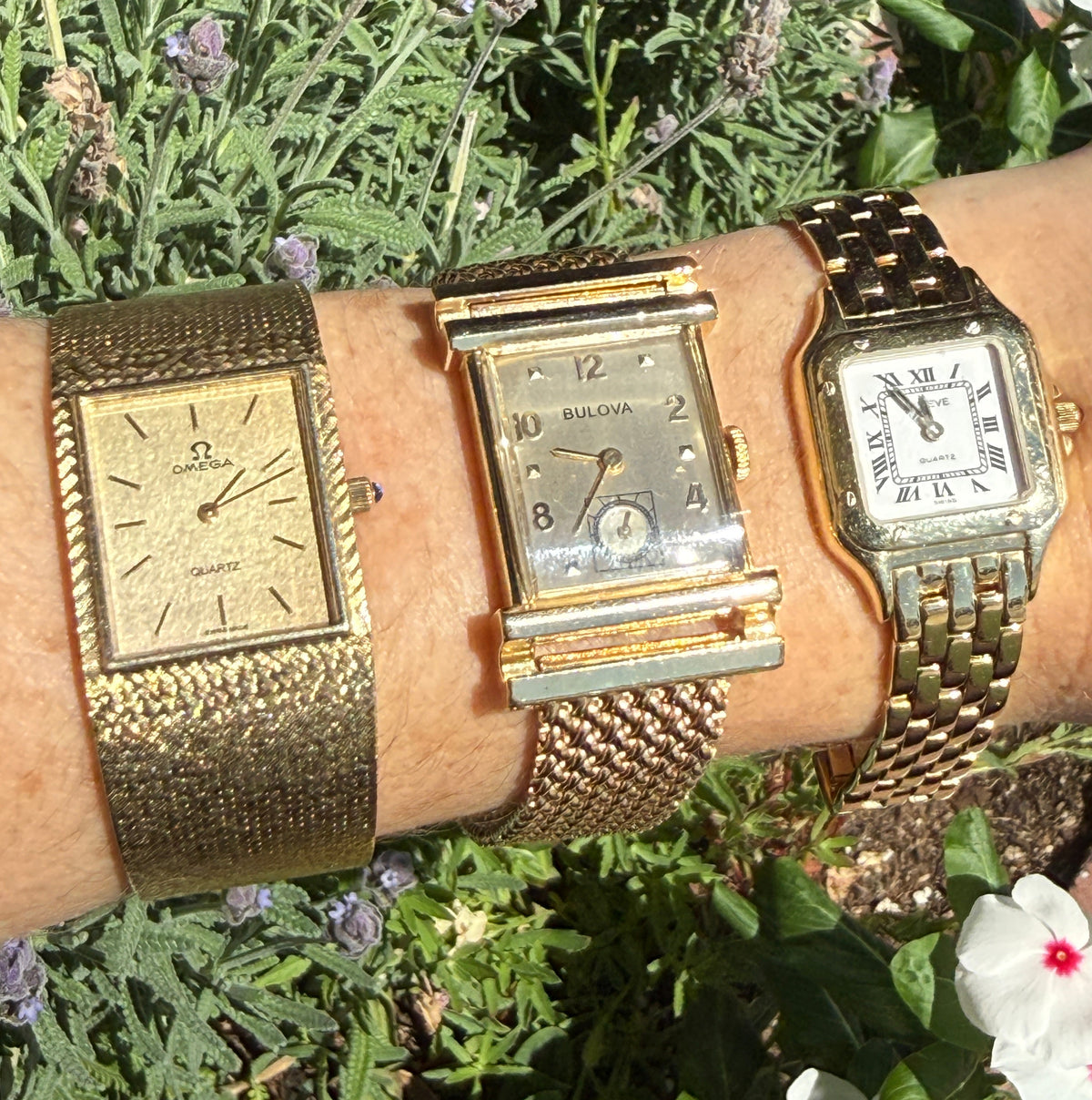 Three vintage-style wristwatches with gold-tone metal cases and mesh bands are stacked on a person's wrist, photographed outdoors against green foliage and small white flowers.